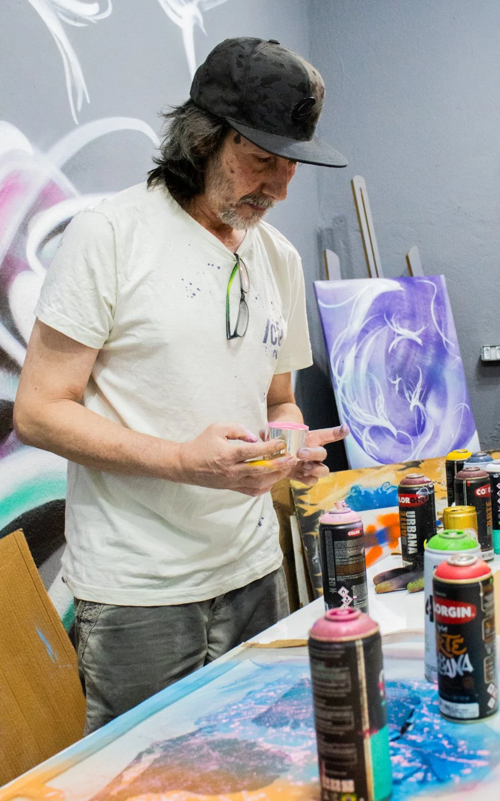 Artista manuseia materiais de pintura diante de uma mesa com sprays e telas