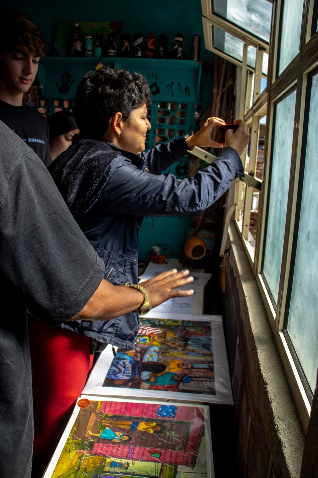 Grupo de visitantes junto à janela, com obras expostas; uma pessoa fotografa o lado de fora
