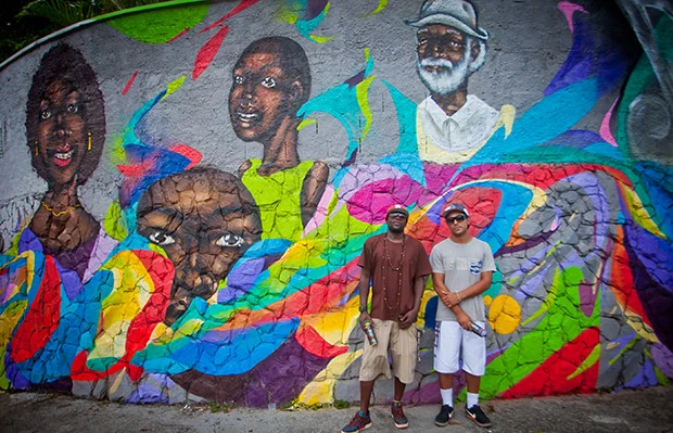 Mural com diversos rostos e duas pessoas diante da pintura