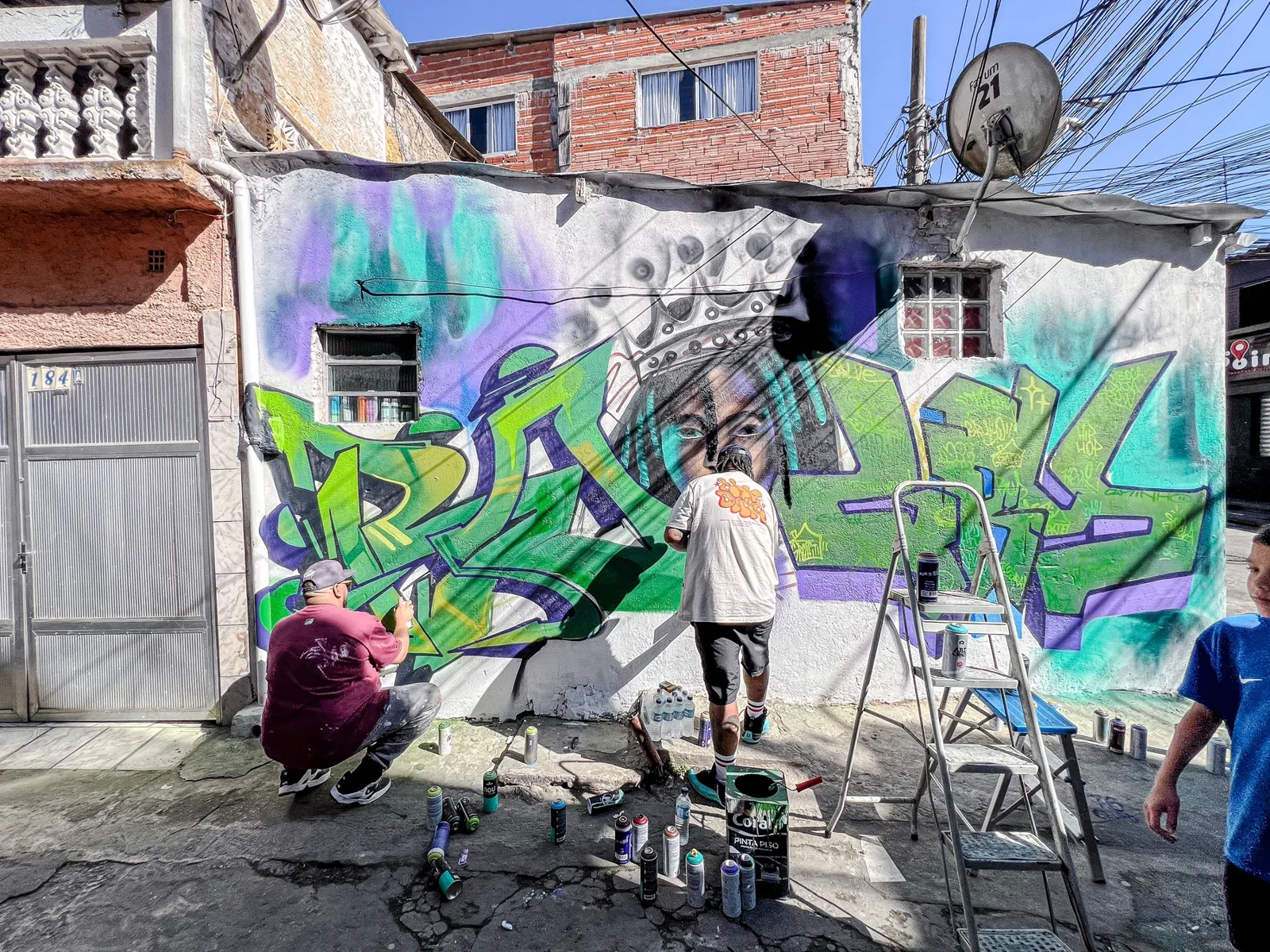 Artistas trabalham em grande pintura de letras verdes e azuis sob fachada residencial
