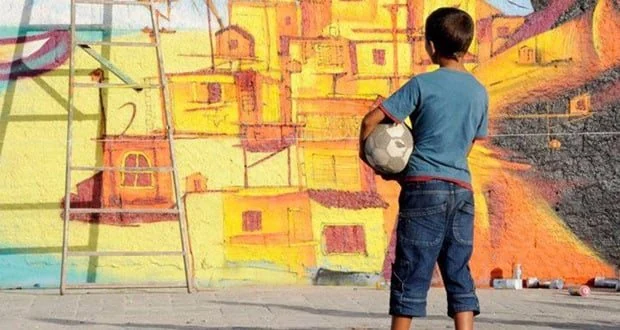 Menino com bola diante de uma pintura de casas e edificações